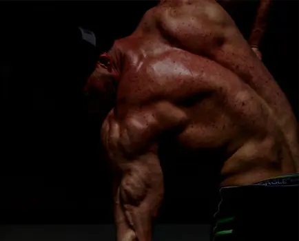 Svalnatý člověk s holým trupem je vyfotografován s dramatickým osvětlením, zvýrazňujícím svaly zad a paží na tmavém pozadí.