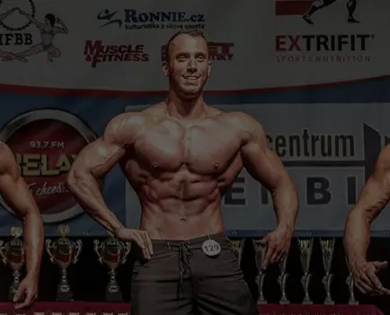 Kulturista stojí na jevišti během soutěže a protahuje svaly s několika trofejemi v pozadí.