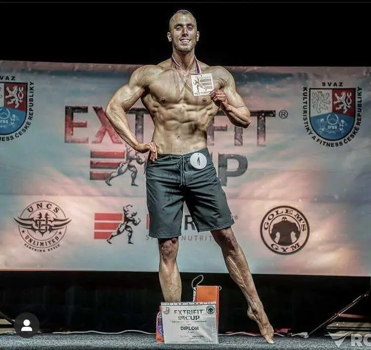 Kulturista pózuje na pódiu před bannerem soutěže fitness, drží certifikát a nosí černé šortky s číslem soutěžícího. Soutěžní loga viditelná na pozadí.