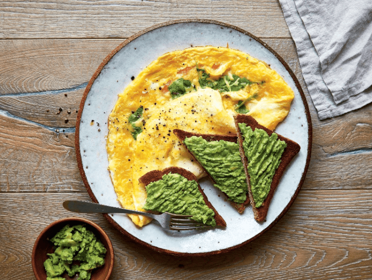 Talíř s omeletou a dvěma plátky toastu s rozmačkaným avokádem, ochucený pepřem, na dřevěném stole. Malá miska s avokádem na boku.