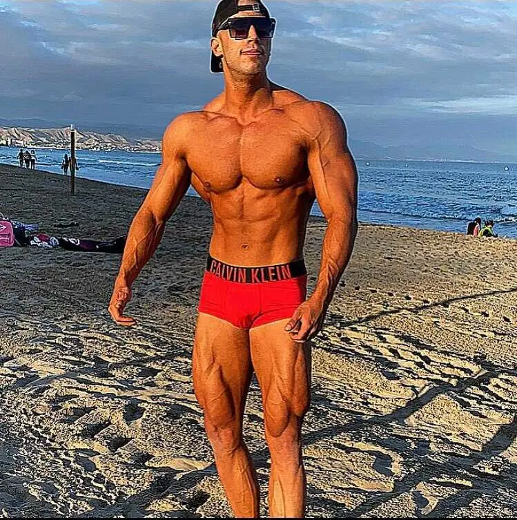 Svalnatý muž v červených šortkách Calvin Klein a slunečních brýlích stojí na písečné pláži s oceánem a zataženou oblohou v pozadí.